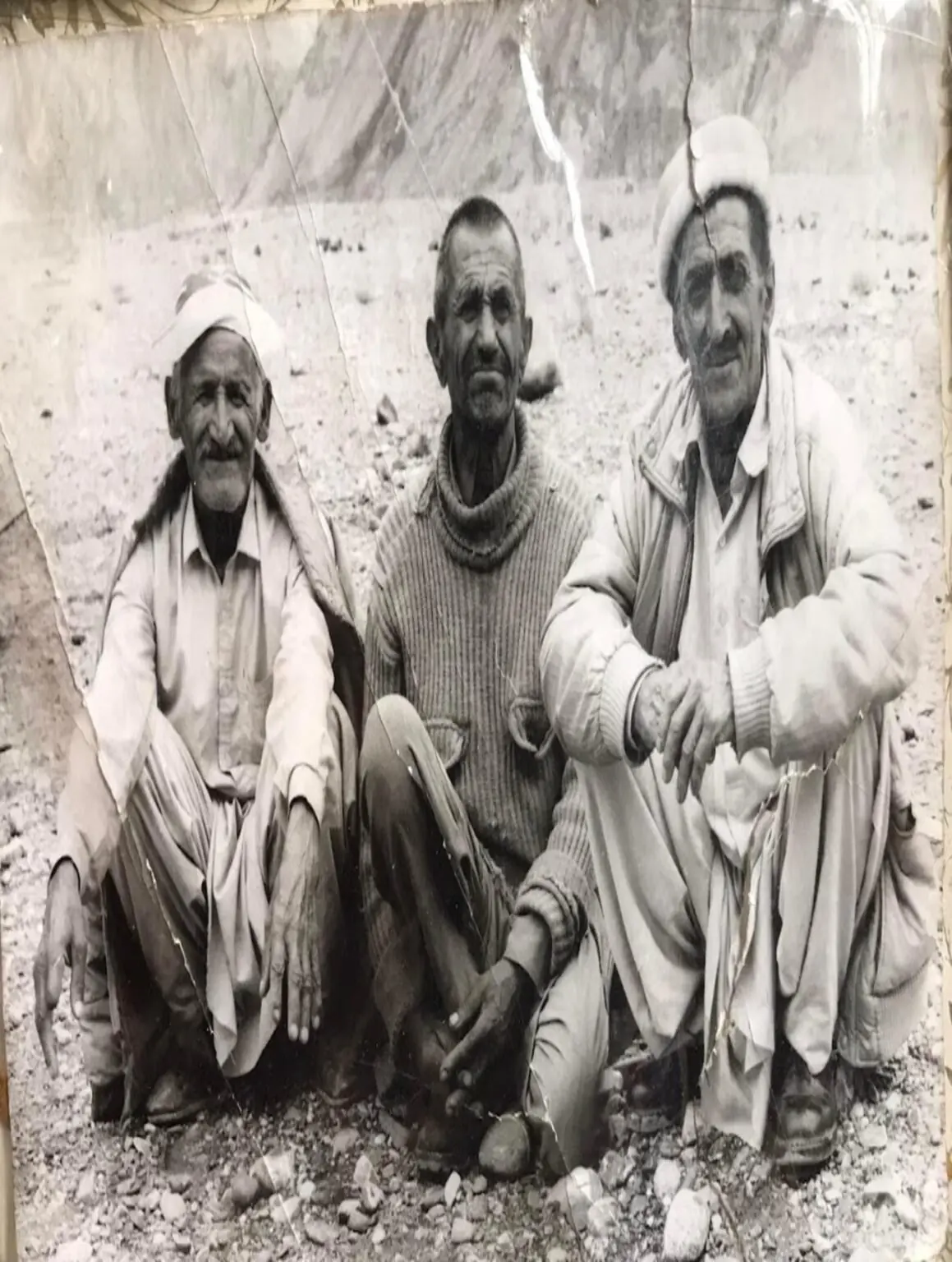 An archival black-and-white photograph of three elders seated on rocky ground
